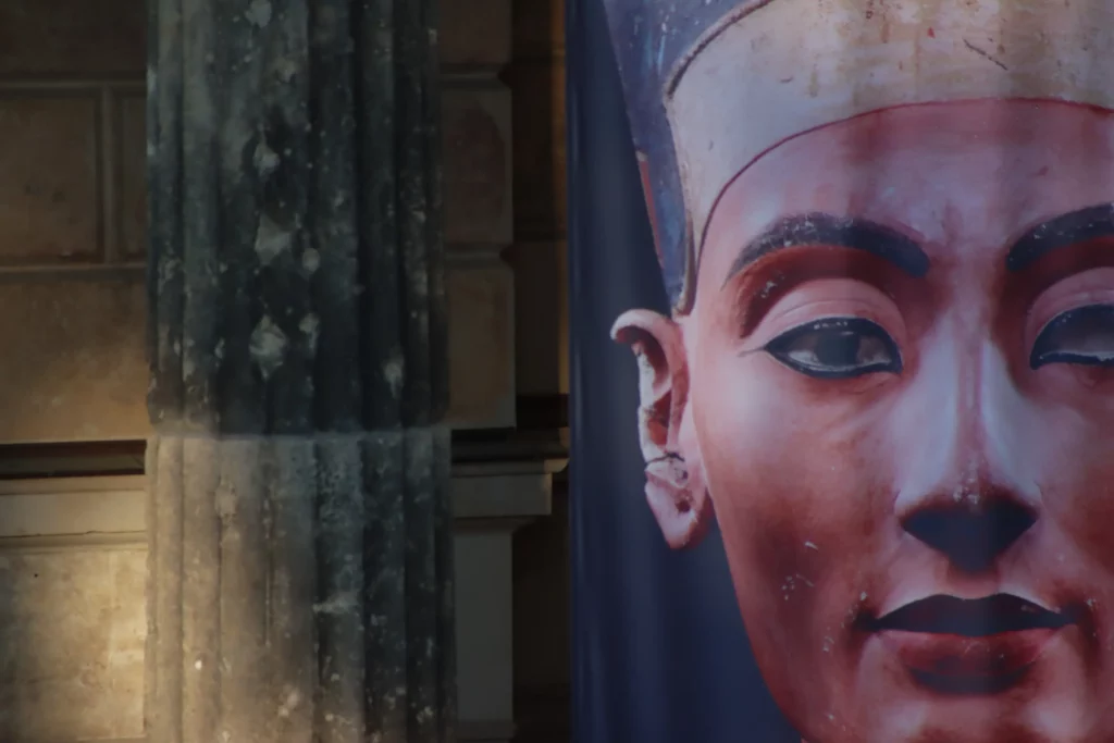 Close-up portrait of Queen Nefertiti's bust on a banner beside a stone column, promoting the Nefertiti Affair outdoor escape game in Berlin